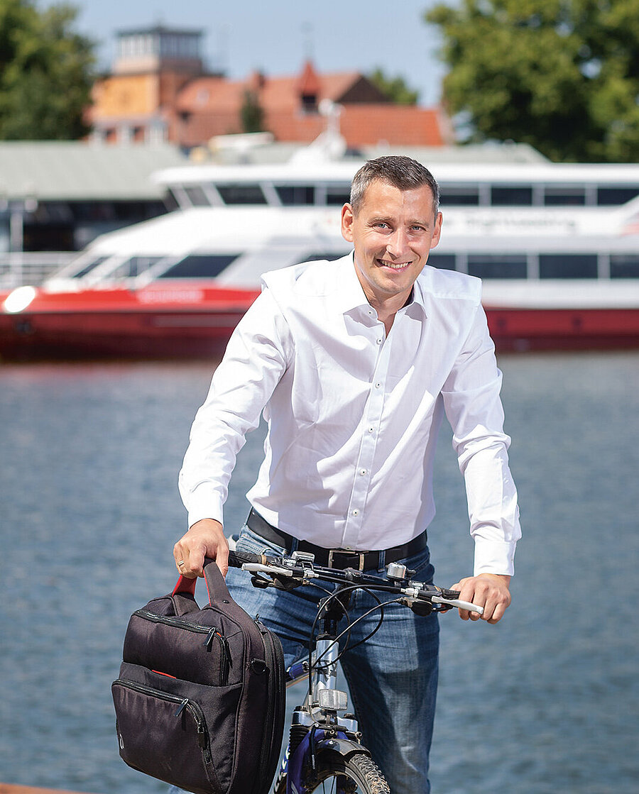 Christian Stolt, Pflegegutachter beim Medizinischen Dienst Mecklenburg- Vorpommern (auf dem Fahrrad, im Hintergrund ein Schiff auf dem Wasser)